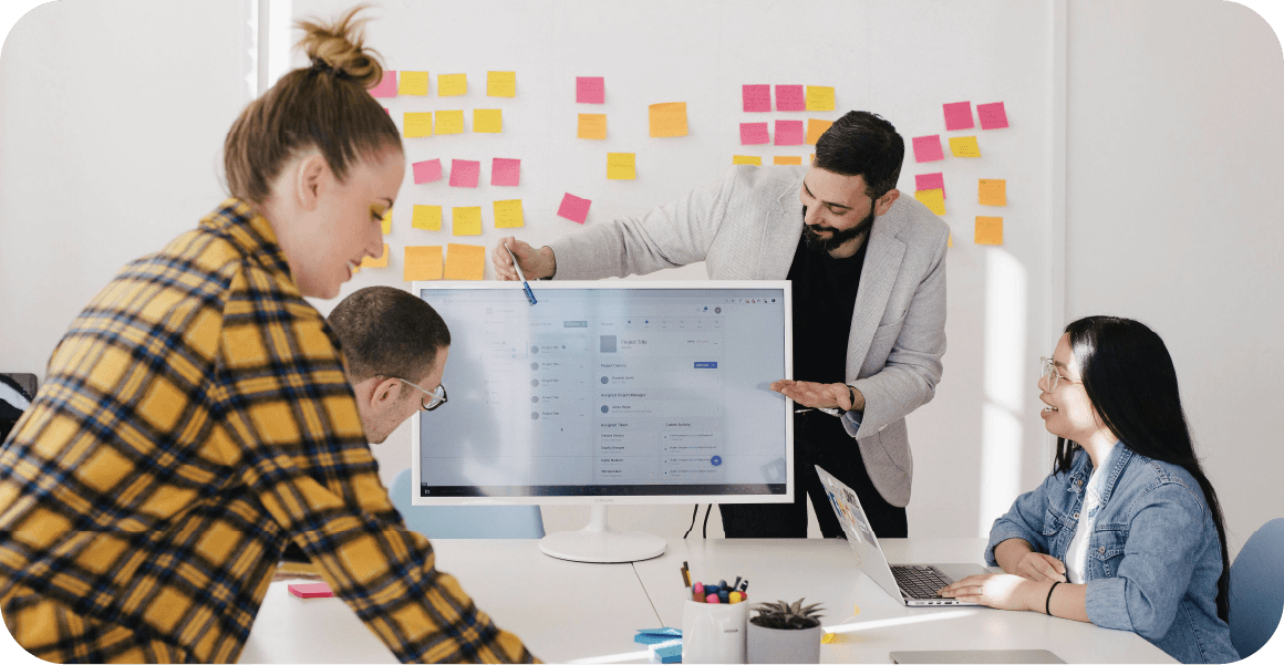 Four people in an office collaborate around a computer screen, with colorful sticky notes on the wall in the background.