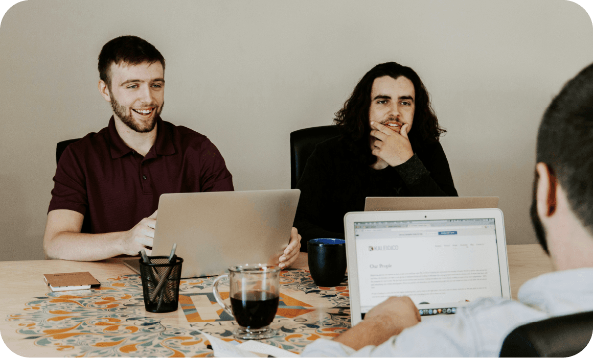 Three people sit at a table with laptops and glasses, engaged in a discussion or meeting.